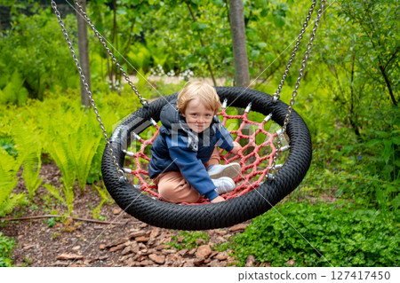 An adorable young child is playing outdoors on a rope swing amidst vibrant greenery, enjoying a delightful moment in nature. An adorable young child is playing outdoors on a rope swing amidst vibrant greenery, enjoying a delightful moment in nature. 127417450