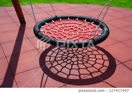 A red net swing casts an intricate shadow on playground tiles, creating visual interest with vibrant colors and geometric shapes in a sunny outdoor park setting. 127417497