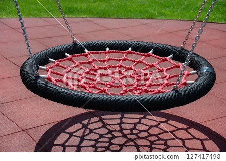 A red net swing casts an intricate shadow on playground tiles, creating visual interest with vibrant colors and geometric shapes in a sunny outdoor park setting. 127417498