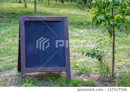 A blank chalkboard sign stands on lush green grass near a young tree in a sunny park. The landscape suggests an invitation for announcements, messages, or creative outdoor presentations. A blank chalkboard sign stands on lush green grass near a young tree in a sunny park. The landscape suggests an invitation for announcements, messages, or creative outdoor presentations. 127417506
