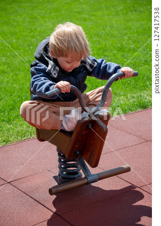 Young child enjoying an outdoor playground, rocking playfully on a wooden rocker. Bright and vibrant setting featuring a safe padded ground and a sunny day, encouraging activity and fun. Young child enjoying an outdoor playground, rocking playfully on a wooden rocker. Bright and vibrant setting featuring a safe padded ground and a sunny day, encouraging activity and fun. 127417538