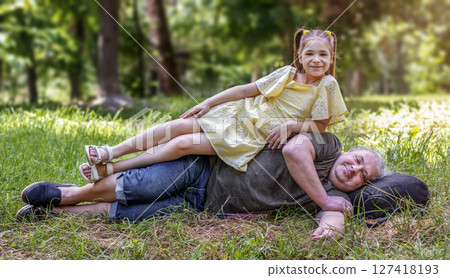 Elderly man lying in grass with child sitting on him in nature Elderly man lying in grass with child sitting on him in nature 127418193