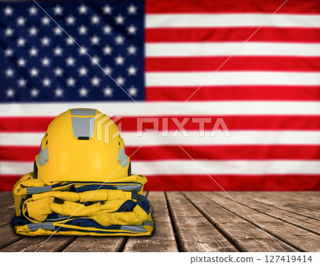 Protective yellow helmet and uniform placed on wooden surface against the background of the United States flag. Stock photo. Protective yellow helmet and uniform placed on wooden surface against the background of the United States flag. Stock photo. 127419414
