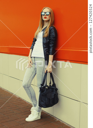 Stylish young woman standing in black leather jacket, sunglasses, modern blonde girl posing in city 127420324