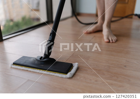 Close-up of unrecognizable man washing hardwood floor with steam cleaner mop in modern apartment, ensuring hygiene and cleanliness. Concept of modern housekeeping appliance. Close-up of unrecognizable man washing hardwood floor with steam cleaner mop in modern apartment, ensuring hygiene and cleanliness. Concept of modern housekeeping appliance. 127420351