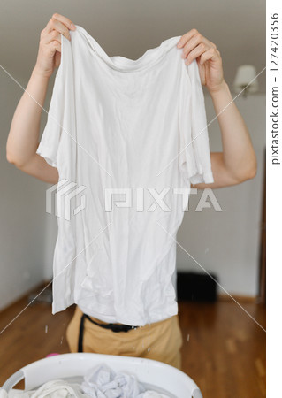 Vertical shot of housekeeper squeezing dripping wet white t-shirt over laundry basket full of soaking clothes, preparing laundry for washing machine, standing in bright living room at home. 127420356