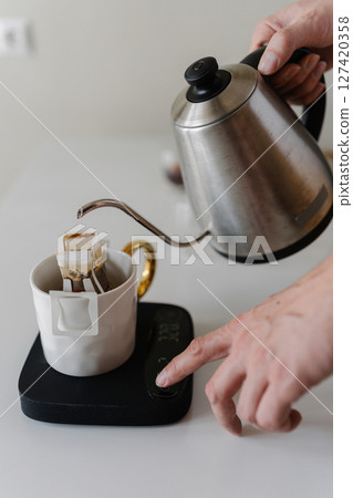 Male hands adjusting kitchen scales during preparing drip coffee, pouring hot water from kettle onto coffee bag on kitchen scales in precise brewing process, close-up, creating morning ritual. 127420358