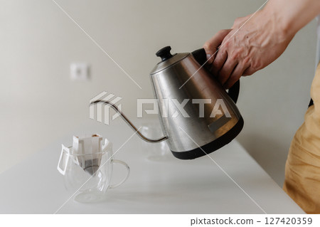 Barista preparing drip coffee bag by pouring hot water from stainless steel kettle into clear glass cup placing on white table at home, creating inviting morning ritual. Barista preparing drip coffee bag by pouring hot water from stainless steel kettle into clear glass cup placing on white table at home, creating inviting morning ritual. 127420359