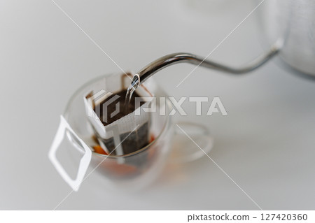 Close-up of hot water pouring from kettle into drip coffee bag placed in glass cup, offering simple and convenient way to brew fresh cup of coffee. Concept of quick way to enjoy fresh cup of coffee. 127420360