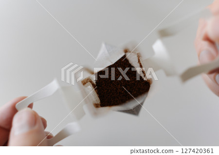 Close-up top view of male hands carefully placing drip coffee bag into transparent glass cup, getting ready to brew fresh cup of coffee in modern kitchen setting at home in morning during breakfast. 127420361