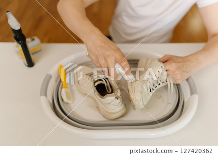 Process of cleaning white dirty sneakers with scrubbing brush and detergent, removing stubborn stains from footwear in white basin, preserving shoe hygiene and appearance thorough washing work. 127420362