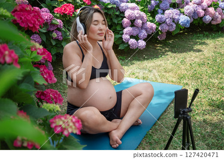 Pregnant woman practicing yoga in a vibrant garden, enjoying music through headphones while recording a video on her smartphone set up on a tripod, embracing relaxation and mindfulness.Music therapy. Pregnant woman practicing yoga in a vibrant garden, enjoying music through headphones while recording a video on her smartphone set up on a tripod, embracing relaxation and mindfulness.Music therapy. 127420471