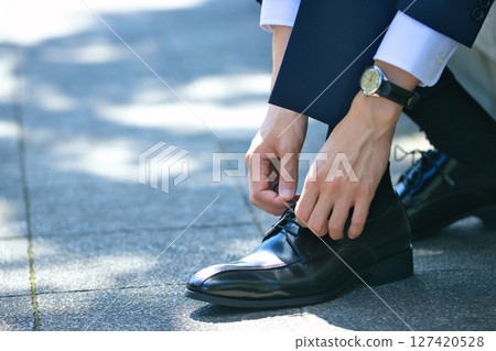 一位穿著西裝的年輕男子正在繫上商務鞋的鞋帶 127420528
