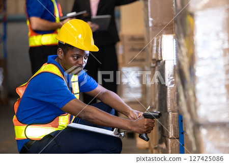 Man African American professional worker wearing safety uniform and white hard hat using bar code reader scanning box inspect product on shelves in warehouse. Man African American professional worker wearing safety uniform and white hard hat using bar code reader scanning box inspect product on shelves in warehouse. 127425086