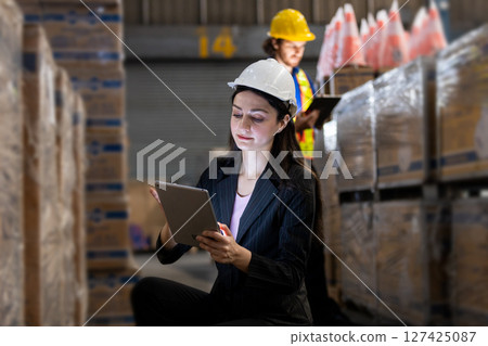 Female in black suit uses digital tablet checking stock goods working in logistics. Women professional warehouse worker wearing safety uniform and white hard hat inspect product at storage. Female in black suit uses digital tablet checking stock goods working in logistics. Women professional warehouse worker wearing safety uniform and white hard hat inspect product at storage. 127425087