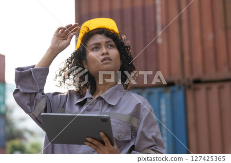 Female African American smiling worker in uniform wear hard hat standing containers yard holding tablet. Area logistics import export and shipping. 127425365