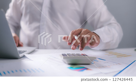 A businessman calculates financial data using a calculator and laptop on a desk filled with charts and reports. Ideal for illustrating accounting, budgeting, finance, and data analysis concepts. 127426445