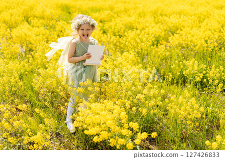 Cheerful child girl with flower crown fooling around and showing tongue looking at camera, holding blank white canvas in vibrant yellow field, offering picturesque setting for mockup designs. Cheerful child girl with flower crown fooling around and showing tongue looking at camera, holding blank white canvas in vibrant yellow field, offering picturesque setting for mockup designs. 127426833