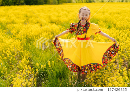 Cheerful young preteen girl in traditional Russian dress posing and smiling in vibrant field of yellow canola flowers on sunny summer day, embracing joy and beauty of childhood in nature landscape. Cheerful young preteen girl in traditional Russian dress posing and smiling in vibrant field of yellow canola flowers on sunny summer day, embracing joy and beauty of childhood in nature landscape. 127426835
