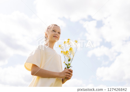 Shooting from below of pretty young girl with braided hair holding bouquet of yellow wildflowers standing posing in sunny summer meadow, enjoying beauty of nature under bright cloudy sky. Shooting from below of pretty young girl with braided hair holding bouquet of yellow wildflowers standing posing in sunny summer meadow, enjoying beauty of nature under bright cloudy sky. 127426842