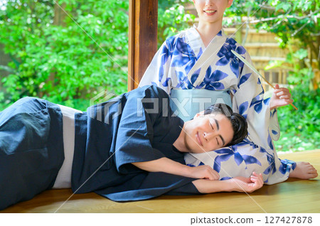 一個溫暖的夏日場景:一位男士微笑著依偎在一位身穿浴衣的女士身邊,在陽台上享受著枕頭放在她腿上的樂趣 一個溫暖的夏日場景:一位男士微笑著依偎在一位身穿浴衣的女士身邊,在陽台上享受著枕頭放在她腿上的樂趣 127427878