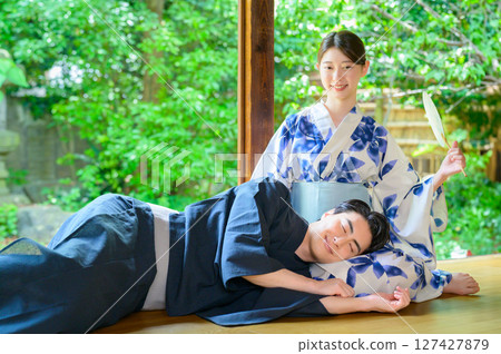 A woman holds a fan and places a man's head on her lap, enjoying the gentle summer sunshine as they relax on the veranda. A woman holds a fan and places a man's head on her lap, enjoying the gentle summer sunshine as they relax on the veranda. 127427879