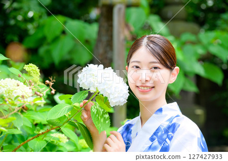 這是一張清新的日式夏日照片，一位身穿浴衣的女子站在寧靜的花園中，手捧白色繡球花，臉上帶著清爽的微笑。 127427933