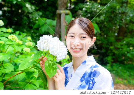 A summer photo of a woman in a yukata with a refreshing smile, holding a white hydrangea in her hands in a garden, blending in with nature. 127427937