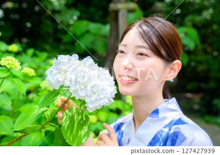 A woman in a yukata smiles as she gazes at the white hydrangeas, enjoying the season in a summer garden enveloped in a serene atmosphere. 127427939