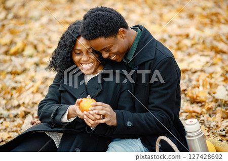 Black man and woman looking on little pumpkin. Romantic couple sitting on a blanket in autumn park. Man and woman wearing black coats. Black man and woman looking on little pumpkin. Romantic couple sitting on a blanket in autumn park. Man and woman wearing black coats. 127428692