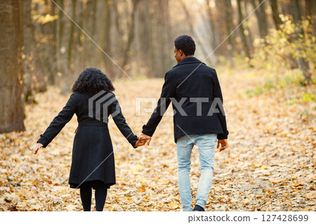 Black young man and his girlfriend holding hands. Romantic couple walking in autumn park. Man and woman wearing black coats. 127428699
