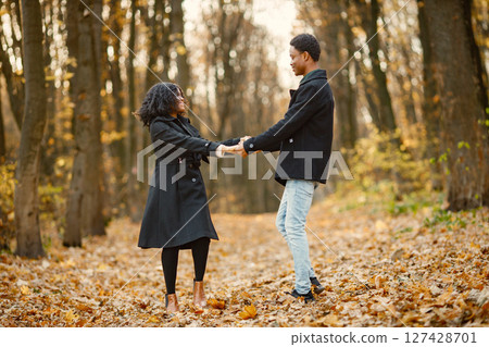 Black young man and his girlfriend dancing. Romantic couple walking in autumn park. Man and woman wearing black coats. 127428701