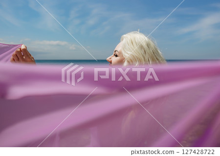Woman beach veil: Blonde woman poses with purple veil on the beach during daytime for art. 127428722
