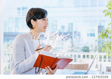 A smiling woman in her 30s interviewing someone in a business setting with her notebook open 127429169