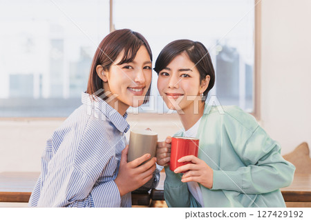 A same-sex couple of women in their 30s looking at the camera, holding mugs in their hands and leaning their faces close together 127429192