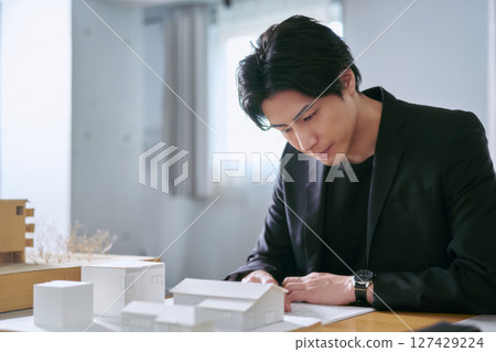 一名男建築師在設計辦公室工作，透過查看建築模型和藍圖來檢查建築藍圖 127429224