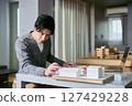 A male student of the Department of Architecture studying the design of a building with a laptop and a drawing in hand while looking at an architectural model 127429228