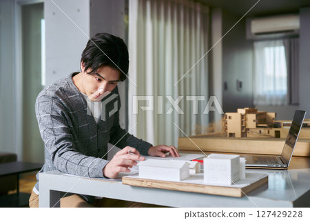 建築系一名男學生手拿筆記型電腦和圖紙，一邊研究建築設計，一邊查看建築模型 127429228