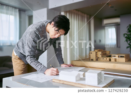 一位男建築師在辦公桌前拿著建築模型檢查和繪製藍圖,同時進行設計 一位男建築師在辦公桌前拿著建築模型檢查和繪製藍圖,同時進行設計 127429230