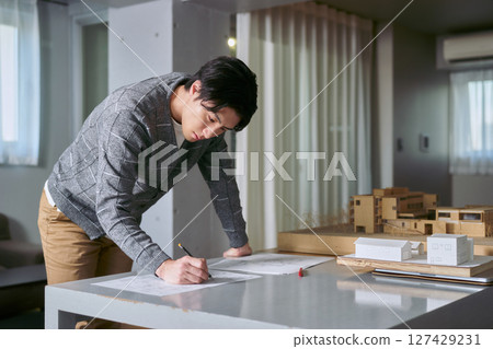 一位在工作室式設計辦公室工作的男建築師站在一張桌子旁,桌上擺放著一棟建築的模型,寫著計劃 一位在工作室式設計辦公室工作的男建築師站在一張桌子旁,桌上擺放著一棟建築的模型,寫著計劃 127429231