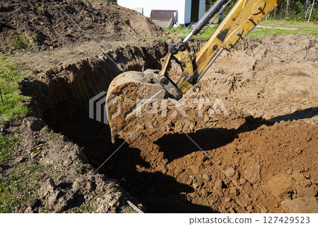A digger operates in a rural setting, carefully excavating a deep trench.  127429523