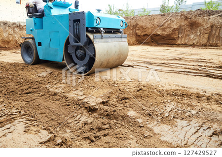 A compact roller is actively compressing dirt on a construction site, where the ground has recently been excavated. A compact roller is actively compressing dirt on a construction site, where the ground has recently been excavated. 127429527