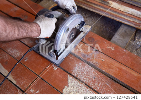 Craftsman operates a circular saw to cut through wooden planks in a well-lit workshop. Craftsman operates a circular saw to cut through wooden planks in a well-lit workshop. 127429541
