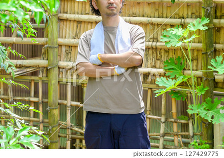 夏天在花園工作時，一名男子在工作休息時雙臂交叉站在竹籬笆前，靜靜地思考下一步該做什麼。 127429775