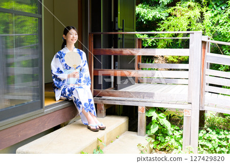 A summer afternoon scene of a woman in a yukata sitting on the veranda of a Japanese-style house, smiling and enjoying the view outside 127429820