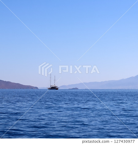 A calm blue sea with a traditional sailing boat in the distance, against a backdrop of hazy mountains and a clear sky. High quality photo 127430977