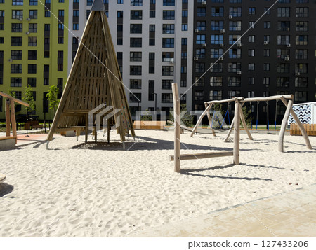 Modern residential playground with sand covered surface, wooden climbing structures, swings and teepee style shelter. Surrounded by colorful apartment buildings and benches in urban setting Modern residential playground with sand covered surface, wooden climbing structures, swings and teepee style shelter. Surrounded by colorful apartment buildings and benches in urban setting 127433206
