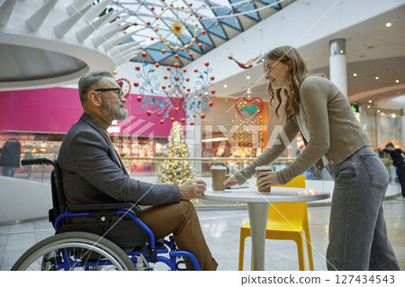 Mature man in wheelchair and young woman-assistant enjoying coffee in a shopping center Mature man in wheelchair and young woman-assistant enjoying coffee in a shopping center 127434543