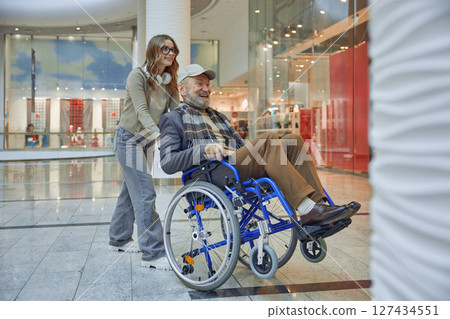 Granddaughter providing assistance to a senior man who uses a wheelchair at a shopping mall 127434551