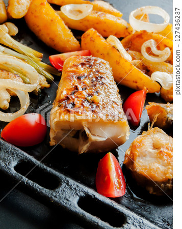 Oven pan with sliced baked potatoes, fish steaks and vegetables with onion on the table. Homemade healthy dinner with potatoes and seafood. 127436287